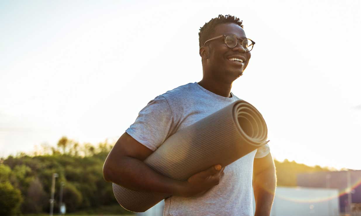 Man with glasses smiling and holding a yoga mat under his arm